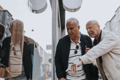 Senior man pointing by elderly male friend while doing window shopping at street