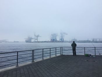 Rear view of man standing on jetty against sea in foggy weather