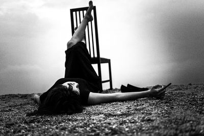 Woman lying on shore by chair against sky