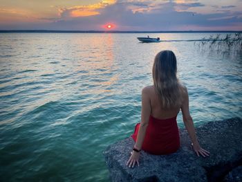 Rear view of woman in red dress sitting down near the lake at sunset and looking at the sky