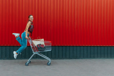 Happy girl with shopping cart on red wall shop background. young woman pushing a shopping cart 