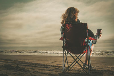 Rear view of woman sitting on beach against sky during sunset