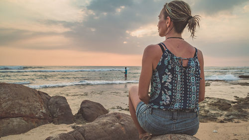 Woman sitting on rock at beach against sky during sunset