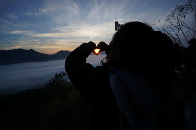 Rear view of silhouette person making heart shape on mountain against sky at sunset