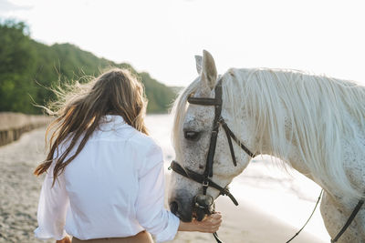 Young long hair woman in white shirt with white horse on seascape background