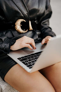 Clever curly businesswoman wearing black leather suit and jacket typing on laptop keyboard while sitting on stairs and working on remote project on city street