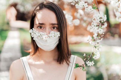 Portrait of sad young woman in protective medical face mask with flowers near blooming tree.