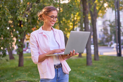 Smiling woman using laptop while standing against trees