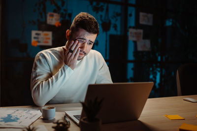 Tired man wearing casual clothes rubbing eyes while working laptop and trying hurry up to deadline