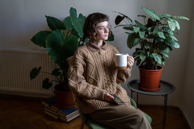 Pensive thoughtful young stylish woman in glasses holding mug looking at window at home with plants.