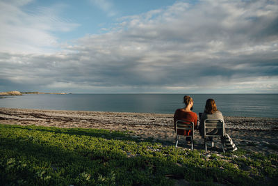 Rear view of friends sitting on chairs at sea shore against cloudy sky