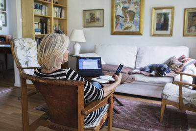 Senior woman using smart phone at home while dog relaxing on sofa