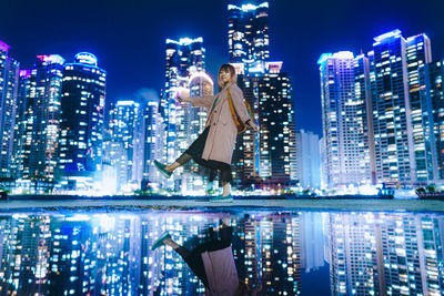 Portrait of woman standing by lake against illuminated buildings in city at night