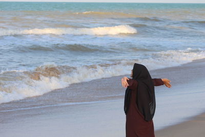 Young woman wearing hijab with arms outstretched standing on shore at beach