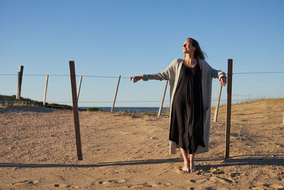 Delighted female in dress standing near fence on sandy beach and enjoying sunny day in summer