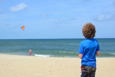 Rear view of boy flying kite with friend at beach against sky