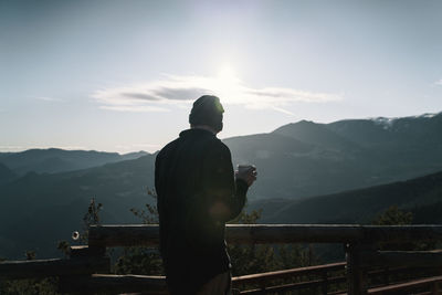 Rear view of man with cup of coffee looking at mountain range against sky