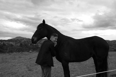 Side view of smiling young man standing with horse on field against cloudy sky