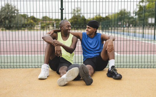 Smiling mature man with son sitting in front of fence at sports court