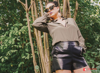 Young woman wearing sunglasses standing by plants against trees