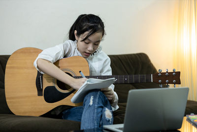 Cute girl writing in notebook while holding guitar guitar at home