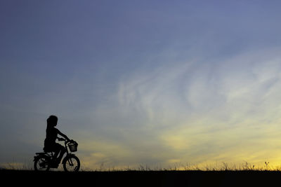 Silhouette girl riding bicycle on field against sky during sunset