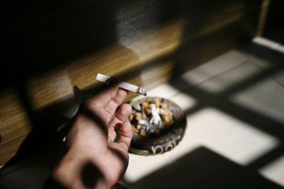 Cropped hand of person holding cigarette by ashtray on floor at home