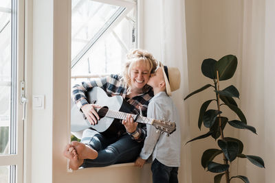 Woman sat playing guitar at home whilst her son gives her a kiss