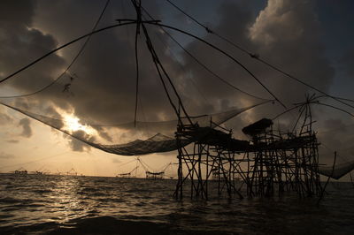 Silhouette chinese fishing nets in sea against cloudy sky