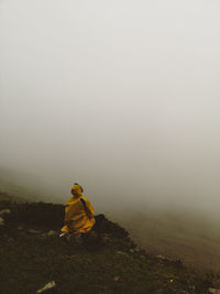 Person sitting on rock against sky during foggy weather