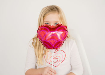 Cute young blond girl holding small red heart balloon under eyes