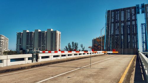 Road by buildings against clear blue sky in city