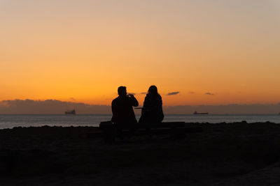 Silhouette couple sitting on beach against orange sky