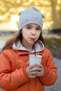 A girl drinks a drink from a straw and a glass, looks at the camera while walking on the street