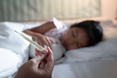 Cropped hand of mother holding thermometer while daughter sleeping on bed