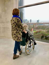 Rear view of two women, one in a wheelchair, in a concrete room looking out over the city