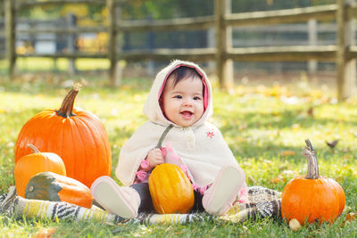 Cute baby girl wearing warm clothing sitting with pumpkins on land