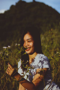 Smiling young woman holding flower while standing on grassy field against mountain