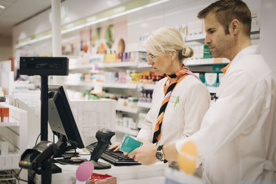 Confident male pharmacist scanning medicine with bar code reader by female colleague using computer at checkout in store