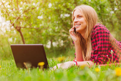 Smiling young woman using laptop while talking on phone outdoors