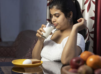 An indian asian woman holding a glass of milk sitting beside a table in a domestic room