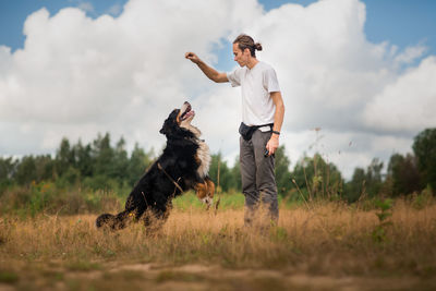 Man playing with dog while standing on grassy land against cloudy sky