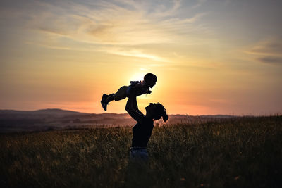 Silhouette mother carrying baby while standing on field against sky during sunset