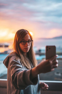 Young woman taking selfie with mobile phone in balcony against sky during sunset