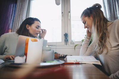 Young female friends studying from book against window in college dorm room