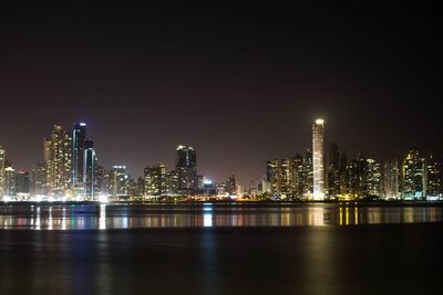 Illuminated buildings by river against sky at night