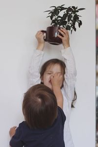 Rear view of boy pinching sister holding houseplant against wall