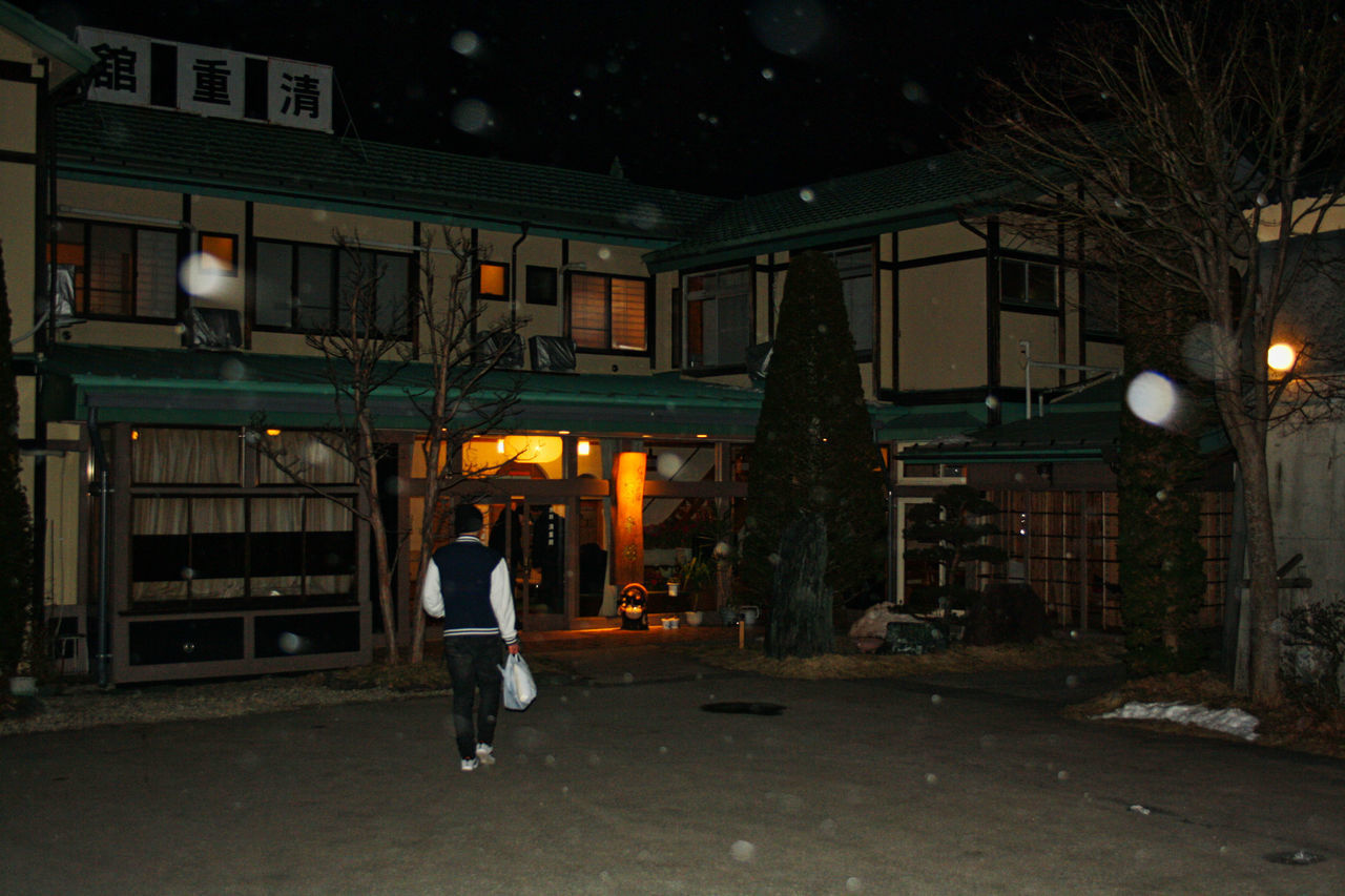 REAR VIEW OF MAN STANDING ON STREET AGAINST ILLUMINATED BUILDINGS