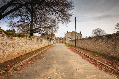 Empty footpath amidst bare trees and buildings against sky