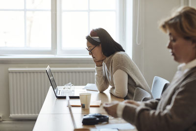 Side view of female teacher watching laptop while sitting by colleague at desk in staffroom